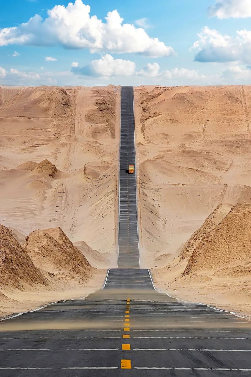 Desert road disappearing into the sandy landscape under a blue sky with clouds.