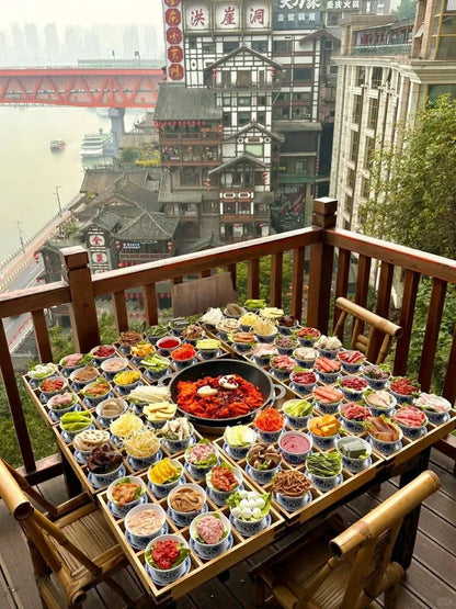 Table overflowing with vibrant dishes showcasing Chongqing's famous hot pot cuisine against a scenic river backdrop.
